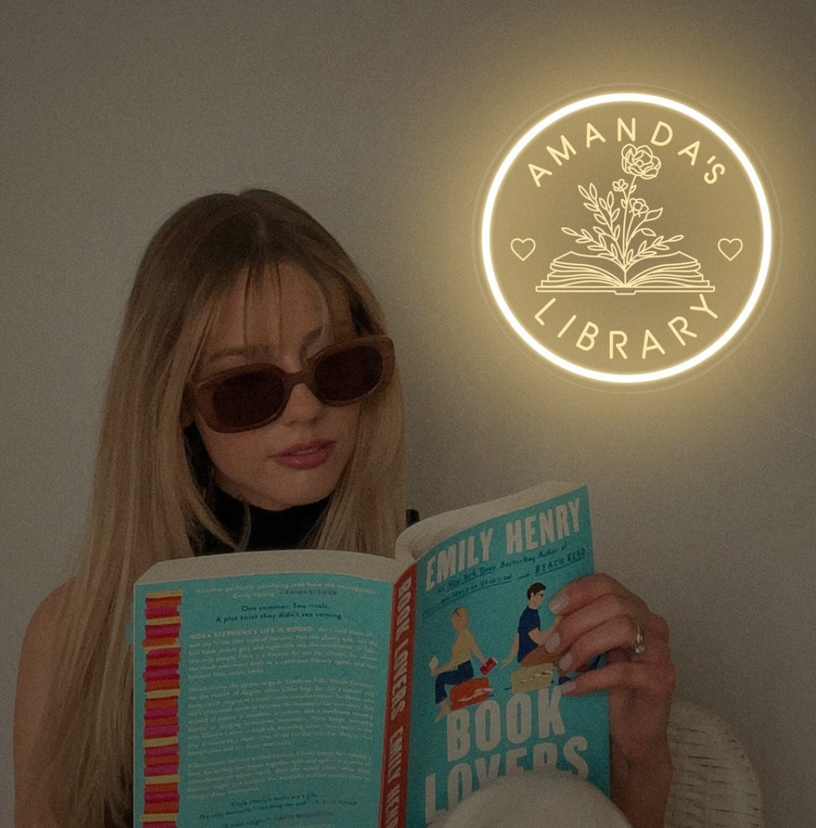 Custom Name's Library Neon Sign on the wall; there is a white woman readng an Emily Henry book in front of it